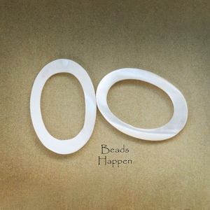 May include: Two white oval-shaped mother of pearl beads. The beads are smooth and have a slight iridescent sheen. The text "Beads Happen" is printed below the beads.