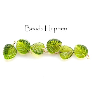 May include: Six translucent green leaf-shaped beads with gold-colored wire loops. The beads have detailed vein patterns and are arranged in a row. The text "Beads Happen" is above the beads.