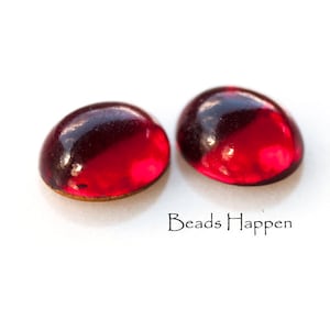 Pode incluir: Duas contas de cabochão de vidro vermelho com acabamento brilhante. As contas são redondas e têm uma ligeira forma de cúpula.
