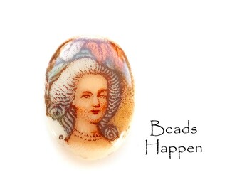 May include: Oval glass bead featuring a painted image of a woman in a white and blue hat. The woman has brown hair and is wearing a white dress with a pearl necklace. The bead has a brown and white background.