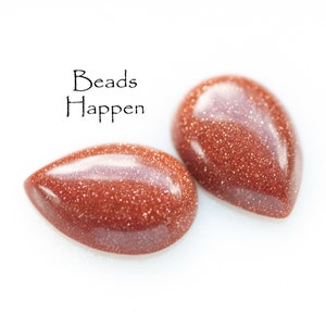 May include: Two teardrop-shaped, reddish-brown gemstone beads with a sparkly, glittery texture. The text "Beads Happen" is written above the beads.