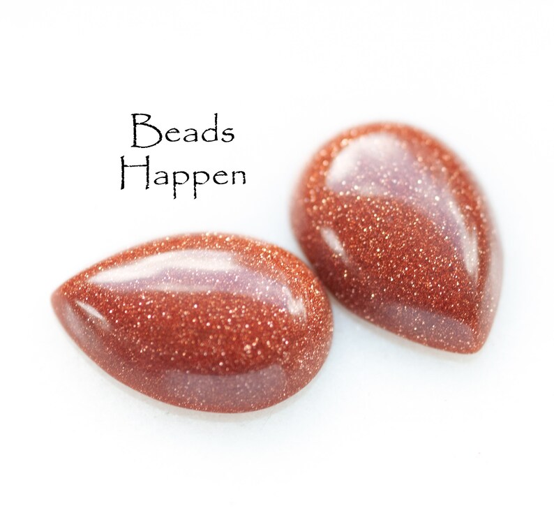 May include: Two teardrop-shaped, reddish-brown gemstone beads with a sparkly, glittery texture. The text "Beads Happen" is written above the beads.