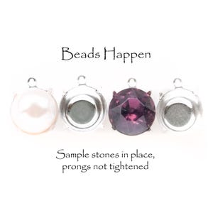 May include: Four silver-toned bezel settings with sample stones. The stones include a white pearl, a gray stone, and a faceted purple stone. The text "Beads Happen" and "Sample stones in place, prongs not tightened" are also visible.