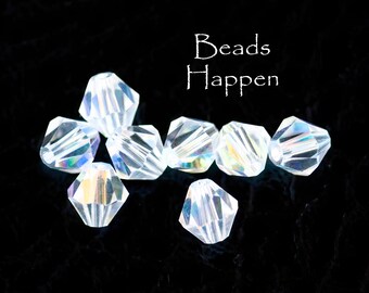 May include: A group of iridescent clear glass beads, with a faceted, teardrop shape. The beads are arranged on a black background. The text "Beads Happen" is written in white above the beads.