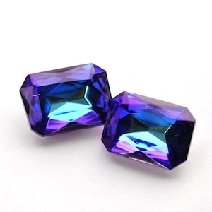 Puede incluir: Dos gemas rectangulares facetadas con un degradado de tonos azules y morados. Las gemas están talladas con múltiples facetas, creando un efecto brillante. Las gemas están sobre un fondo blanco.
