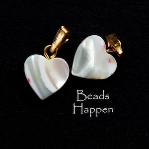 May include: Two white heart-shaped pendants with gold accents. The pendants have a pearlescent finish and a subtle red stripe. The text "Beads Happen" is written below the pendants.