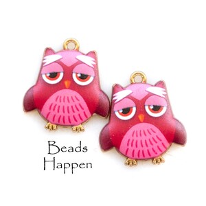 May include: Two red and pink owl charms with gold-colored accents. The owls have a sad expression with large red eyes and a white brow. The text "Beads Happen" is below the charms.