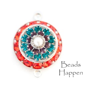 May include: A round, decorative bead with a silver-colored metal base. The bead features a central pearl surrounded by teal and purple gemstones, with a ring of red gemstones around the outer edge. The text "Beads Happen" is in the lower right corner.