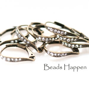 May include: A group of antique gold-toned earring findings with clear rhinestone accents. The findings are shaped like a teardrop and have a hinged closure. The findings are arranged in a pile on a white background. Beads Happen is printed in black text below the findings.