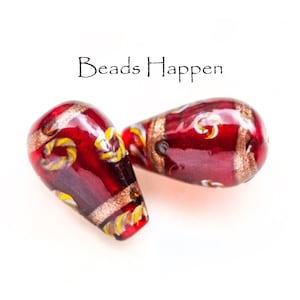 May include: Two red glass beads with gold and white accents. The beads are teardrop shaped and have a glossy finish. "Beads Happen" is written in black text above the beads.