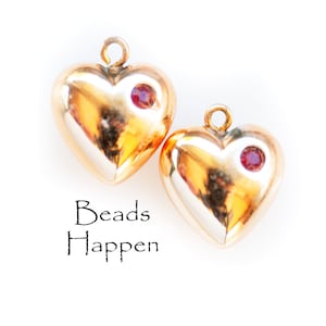 May include: Two gold heart-shaped charms with red gemstone accents. The charms have a shiny, polished finish and are perfect for jewellery making.