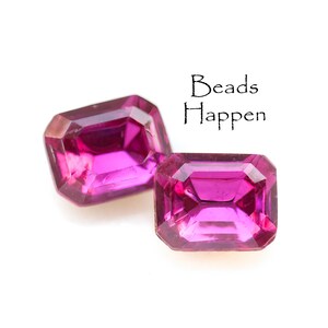 May include: Two pink emerald-cut glass beads. The beads are faceted and have a high shine. The text "Beads Happen" is visible in the background.
