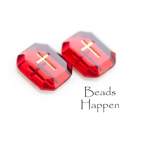 May include: Two red glass beads with gold cross accents. The beads are shaped like a rectangle with rounded corners. The text "Beads Happen" is printed below the beads.