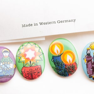 May include: Four oval-shaped Christmas-themed pins. The first pin features a purple background with a depiction of Mary, Joseph, and baby Jesus. The second pin has a green background with a red and yellow star. The third pin has a green background with two yellow candles. The fourth pin has a blue background with three wise men in colorful robes.