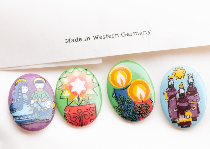 May include: Four oval-shaped Christmas-themed pins. The first pin features a purple background with a depiction of Mary, Joseph, and baby Jesus. The second pin has a green background with a red and yellow star. The third pin has a green background with two yellow candles. The fourth pin has a blue background with three wise men in colorful robes.
