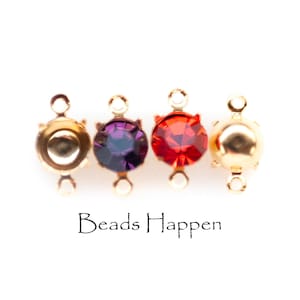 May include: Four gold-colored jewelry findings with colorful crystal accents. One is empty, one has a purple crystal, one has a red crystal, and one has a gold-colored sphere. The text "Beads Happen" is below the findings.