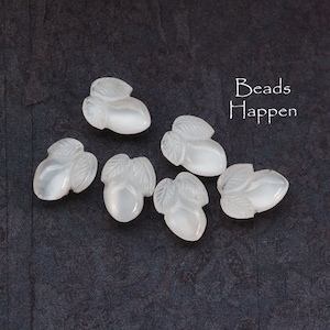 May include: Seven white opaque glass beads with a carved acorn design. The beads are on a dark gray background. The text "Beads Happen" is in white text on the top right of the image.