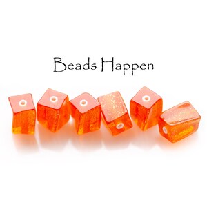 May include: Six orange, cube-shaped beads with a glittery effect and white holes. The beads are arranged in a row against a white background. The text "Beads Happen" is above the beads.
