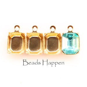 Könnte beinhalten: Vier goldfarbene Metallfassungen mit einem einzigen türkisfarbenen Edelstein in der letzten Fassung. Die Fassungen haben eine quadratische Form mit abgerundeten Ecken und eine Schlaufe oben. Der Text "Beads Happen" ist unter den Fassungen gedruckt.