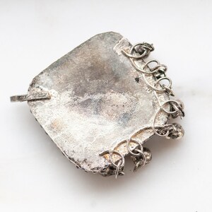 May include: A vintage silver pendant with a square shape and a textured surface. The pendant has a small loop on one side and a decorative chain attached to the bottom. The metal has a tarnished appearance, suggesting age.