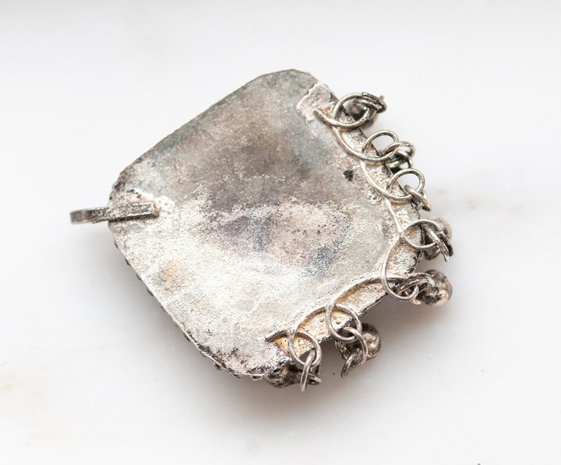May include: A vintage silver pendant with a square shape and a textured surface. The pendant has a small loop on one side and a decorative chain attached to the bottom. The metal has a tarnished appearance, suggesting age.