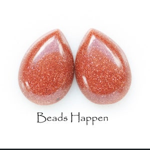 May include: Two teardrop-shaped beads made of red sandstone with a glittery finish. The beads are about 1 inch long and 1/2 inch wide. Beads Happen.