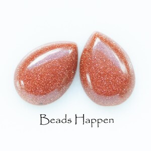 May include: Two teardrop-shaped beads made of red sandstone with a glittery finish. The beads are on a white background with the text "Beads Happen" below them.