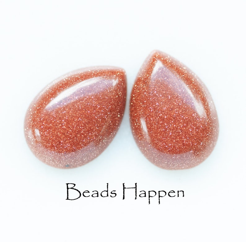 May include: Two teardrop-shaped beads made of red sandstone with a glittery finish. The beads are on a white background with the text "Beads Happen" below them.