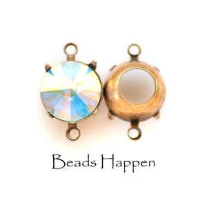 May include: Two antique bronze-colored jewelry components. One features a large, iridescent, round crystal held by prongs. The other is a matching setting with a large, open center. The text "Beads Happen" is below the components.