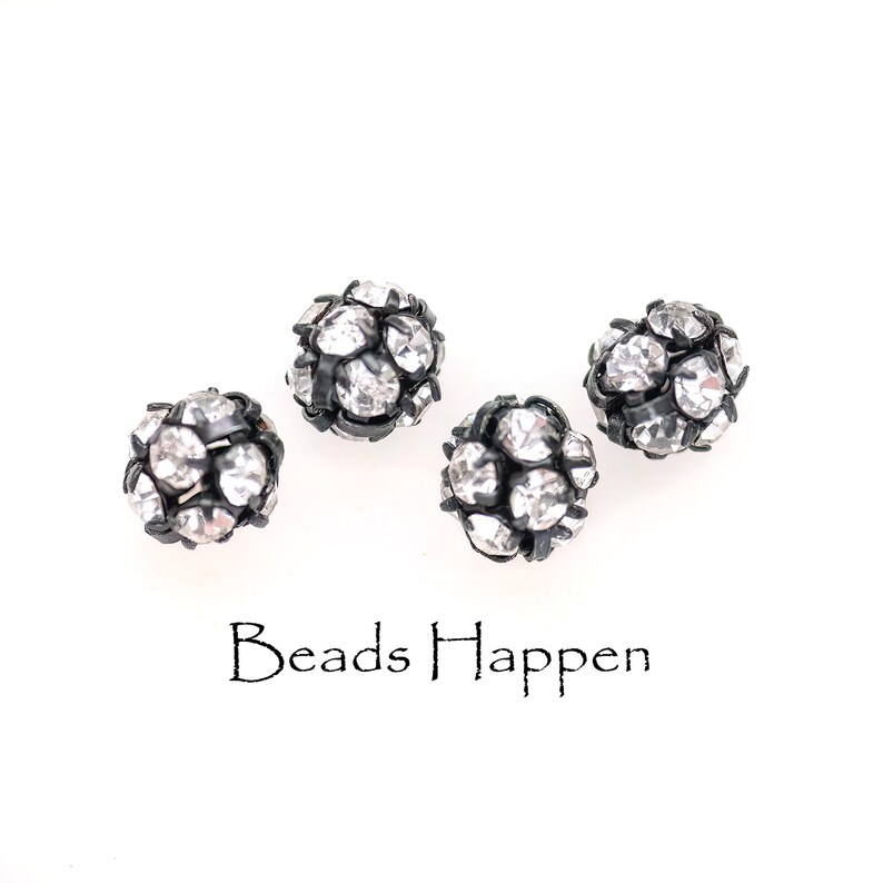 May include: Four black and silver round beads with rhinestones. The beads are scattered on a white surface. The text "Beads Happen" is printed below the beads.