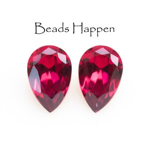 May include: Two faceted, teardrop-shaped, deep red gemstones. The gems are displayed against a white background. The text "Beads Happen" is above the gemstones.