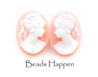May include: Two oval-shaped cameo pendants. Each pendant features a white profile of a woman against a peach-colored background. The text "Beads Happen" is printed in black below the pendants.