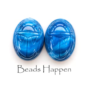 May include: Two blue scarab beetle shaped beads with a white swirl pattern. The beads are on a white background with the text "Beads Happen" below them.