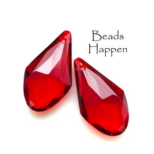 May include: Two faceted, teardrop-shaped, red crystal beads. Each bead has a hole at the top for stringing. The text "Beads Happen" is in black script on a white background.