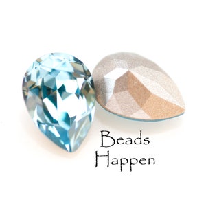 May include: Two pear-shaped, faceted crystal beads. One is a light blue color, and the other is a neutral, metallic tone. The text "Beads Happen" is written in black script below the beads.