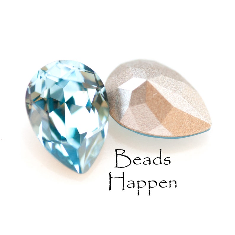 May include: Two pear-shaped, faceted crystal beads. One is a light blue color, and the other is a neutral, metallic tone. The text "Beads Happen" is written in black script below the beads.