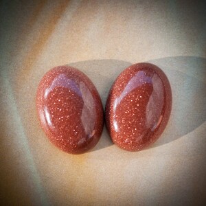 Puede incluir: Dos cabujones ovalados de arenisca pulida. Las piedras son de color marr&oacute;n rojizo intenso con un brillo sutil.