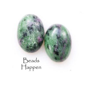 May include: Two green, black, and purple oval-shaped gemstone beads. The beads are smooth and have a speckled pattern. The text "Beads Happen" is written below the beads.