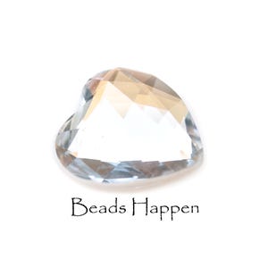 May include: A clear glass heart-shaped bead with a faceted design. The bead is on a white background. Beads Happen.