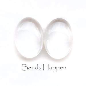 May include: Two clear glass oval cabochons, each with a smooth, polished surface. The cabochons are approximately 2.5 cm in length and 2 cm in width. The cabochons are on a white background. The text "Beads Happen" is printed below the cabochons.