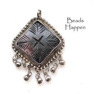 May include: A silver-toned pendant with a diamond-shaped, dark gray center featuring etched lines and a central cross design. The pendant is framed with a beaded border and dangling bell charms. The text "Beads Happen" is visible.