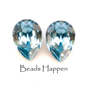 May include: Two faceted, teardrop-shaped, light blue crystals. The crystals are displayed against a white background. The text "Beads Happen" is written in a cursive font below the crystals.