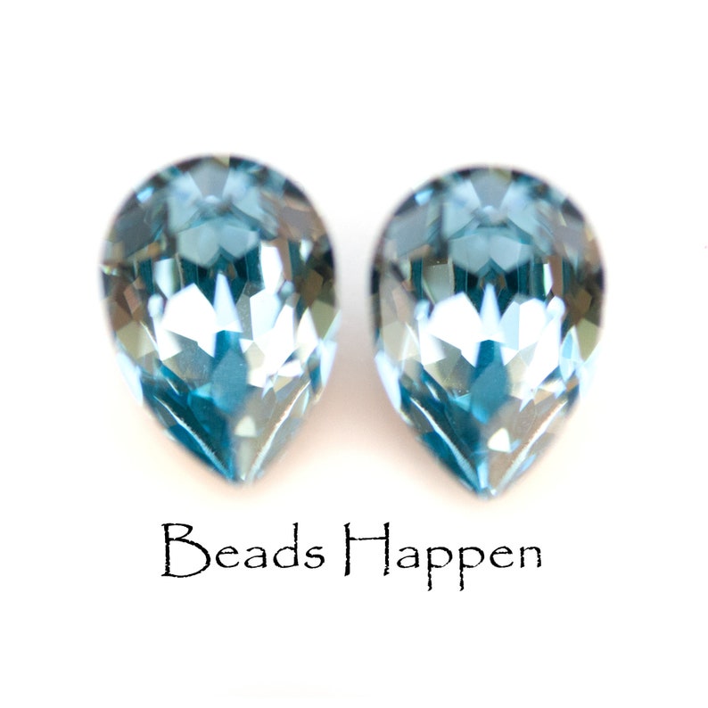 May include: Two faceted, teardrop-shaped, light blue crystals. The crystals are displayed against a white background. The text "Beads Happen" is written in a cursive font below the crystals.