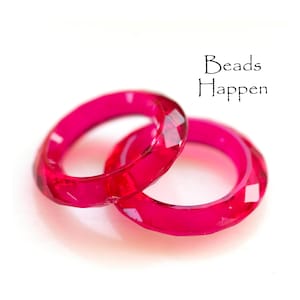 May include: Two faceted, bright pink rings are displayed on a white background. The rings are made of a translucent material and have a geometric design. The text "Beads Happen" is written in black script.