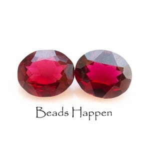 May include: Two oval-shaped, faceted, red gemstones. The gemstones are set against a white background.