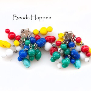 Pode incluir: Um par de brincos pendentes coloridos com uma mistura de contas vermelhas, amarelas, azuis, verdes e brancas. Os brincos são presos a um fecho de clipe prateado. O texto "Beads Happen" é visível acima dos brincos.