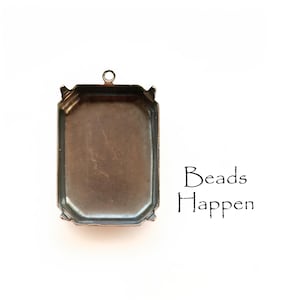 May include: A bronze-colored metal pendant setting with a rectangular shape and a beveled edge. The setting has a loop at the top for attaching a chain or cord. The text "Beads Happen" is printed below the setting.