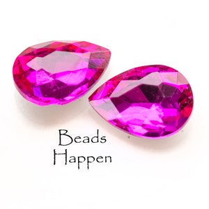 Puede incluir: Dos cuentas de cristal rosa en forma de l&aacute;grima. Las cuentas son facetadas y tienen un alto brillo. El texto "Beads Happen" est&aacute; impreso debajo de las cuentas.