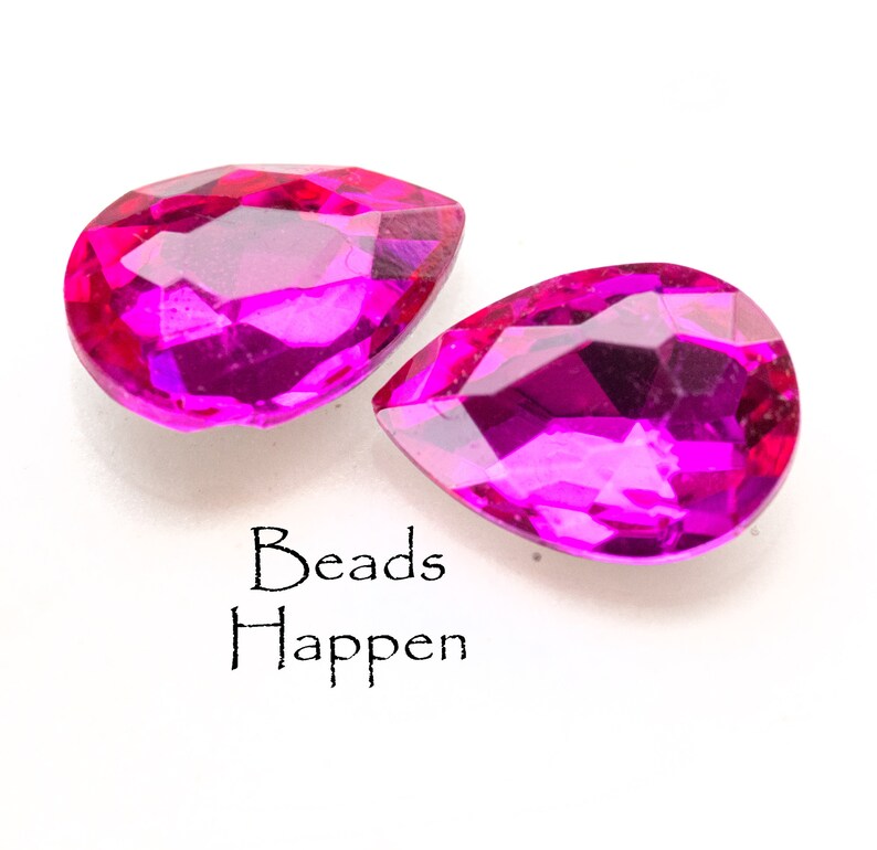 Puede incluir: Dos cuentas de cristal rosa en forma de l&aacute;grima. Las cuentas son facetadas y tienen un alto brillo. El texto "Beads Happen" est&aacute; impreso debajo de las cuentas.