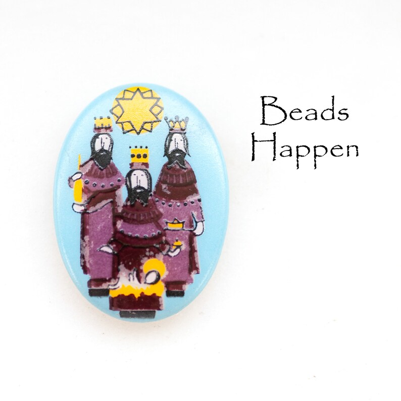 May include: A blue oval-shaped Christmas ornament with a depiction of the three wise men and a star. The ornament has the words "Beads Happen" printed on it.